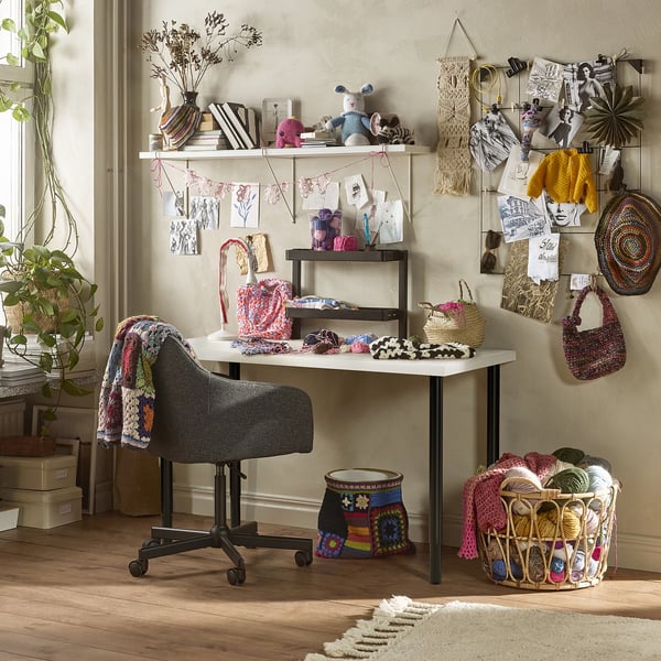 LAGKAPTEN desk with yarn, baskets, & knitting tools. Grey chair with crocheted blanket. Wall adorned with macramé & knitted crafts.
