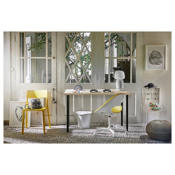 Modern office setup with yellow and grey furniture. Wooden desk, white rubbish bin, and abstract artwork. Large window with plants.