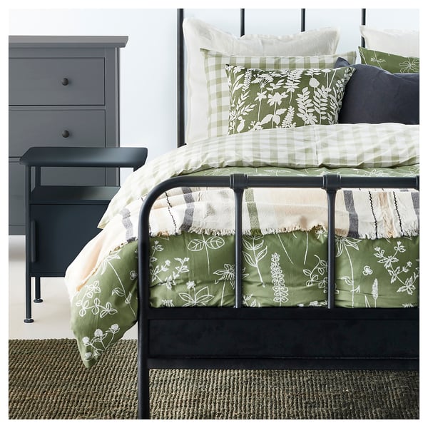 A cosy bedroom with a green and white bedding set and a grey dresser. A dark green jute rug is on the floor.