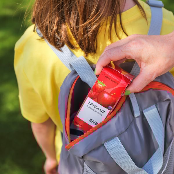 Person holding LÅNGLUR fruit smoothie carton, wearing a yellow shirt and grey backpack.