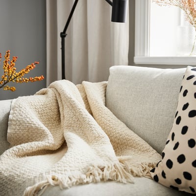 Off-white LAPPTÅG throw blanket, soft & warm, on sofa with black-spotted cushion.