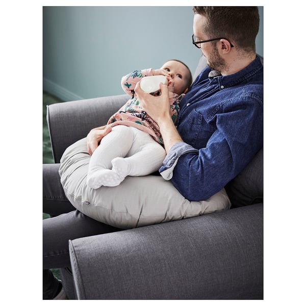 A person holds a baby with a nursing pillow on a grey sofa. The pillow is white and cushions the baby for feeding.