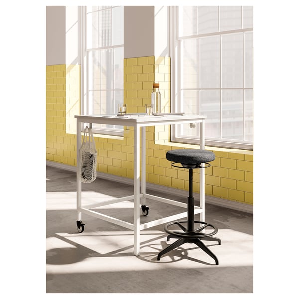 Modern desk with rolling black stool. White metal frame, grey seat. Yellow-tiled wall, windows behind.