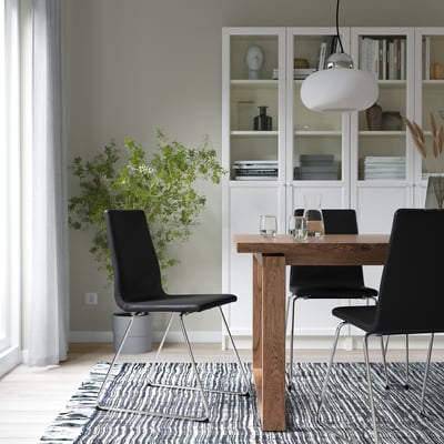 Modern dining room with black chairs, wooden table, white pendant lamp, grey rug, bookshelf, and plant.