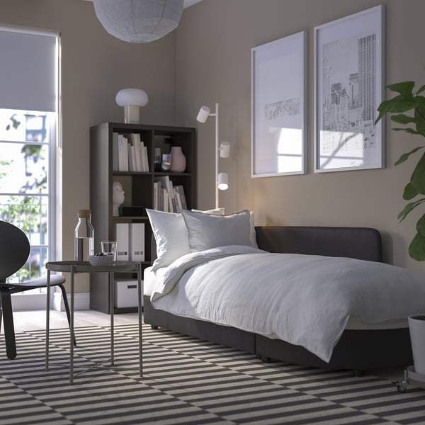 Modern bedroom with grey sofa bed, black and white striped rug, tall bookcase with decor, and large windows.