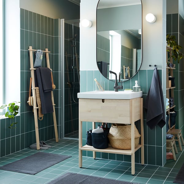 Modern bathroom with green tiles, light wood vanity, round mirror, black fixtures, woven baskets, and dark towels.