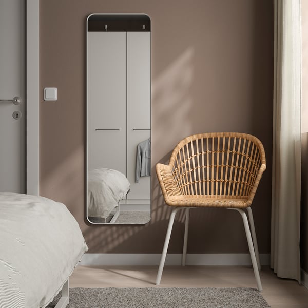 A LINDBYN mirror with white frame, rounded corners, reflecting a bed and wardrobe. Beside it, a white woven chair.