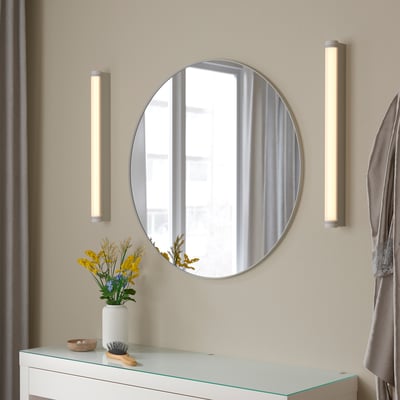 Round lindby mirror with white frame & lights. Below, flower vase & brush on glass chest.