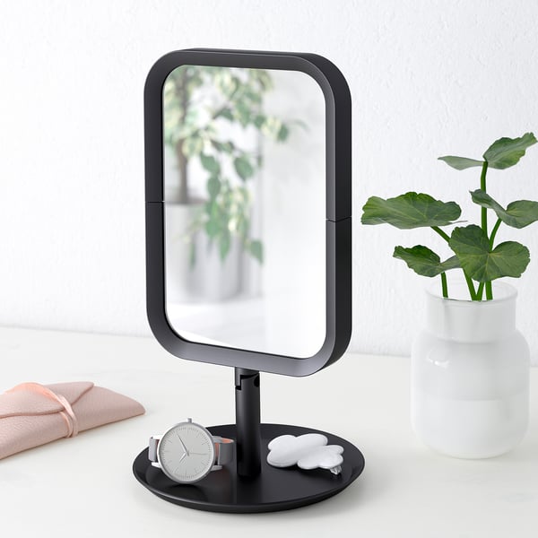 Modern vanity mirror with magnifying side, placed on a table with accessories and a plant.