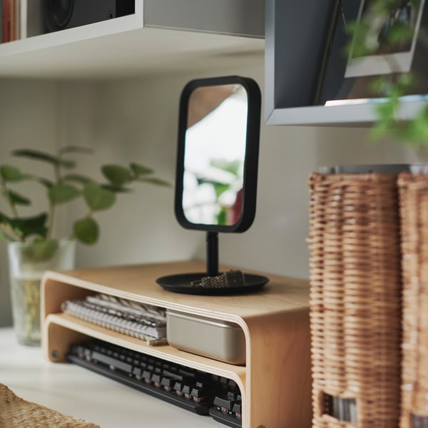 Small black LINDBYN makeup mirror on wooden desk with plant and basket.