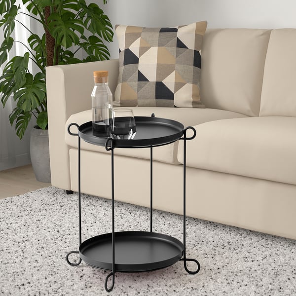 Black side table with two round trays and loop handles, fitting beside sofa and near a large potted plant.
