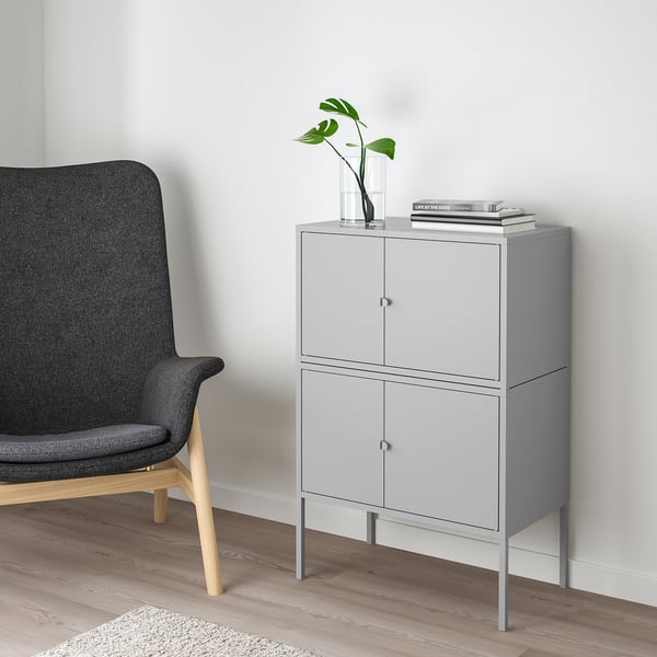 Grey LIXHULT cabinet with 2 doors, slim legs. Beside a dark chair, it holds a plant vase and books.