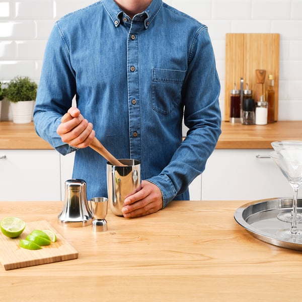 Person in denim shirt uses a cocktail shaker set to prepare drinks on a wooden counter. Includes a shaker, jigger, and cutting board with limes.