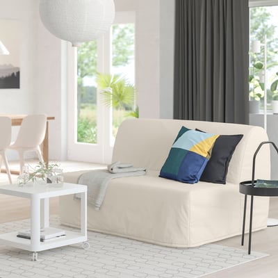 Modern living room with beige lycksele sofa-bed, colourful pillows, white coffee table, grey rug, large windows.
