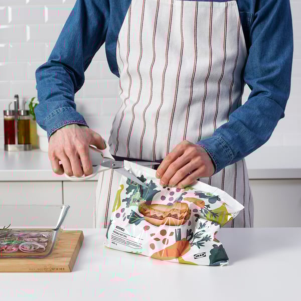 Person in denim shirt and apron opens bag with MÄRKBART scissors, showing its multi-use functionality in the kitchen.