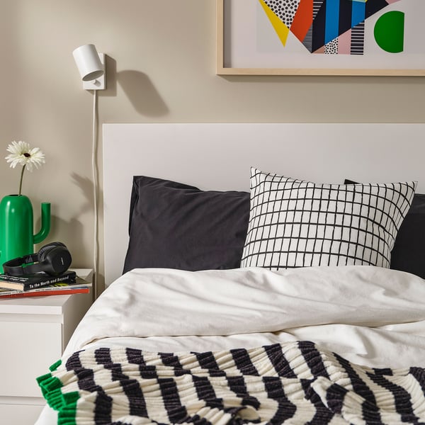 Bed with black & white chequered pillow, green vase, white lamp, headphones, books, abstract wall art.