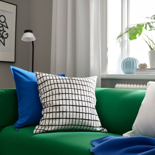 Bright living room with green sofa, blue pillows, chequered cushion, white lamp, and plants. MÄSTERROT pillow in black/white grid is prominent.