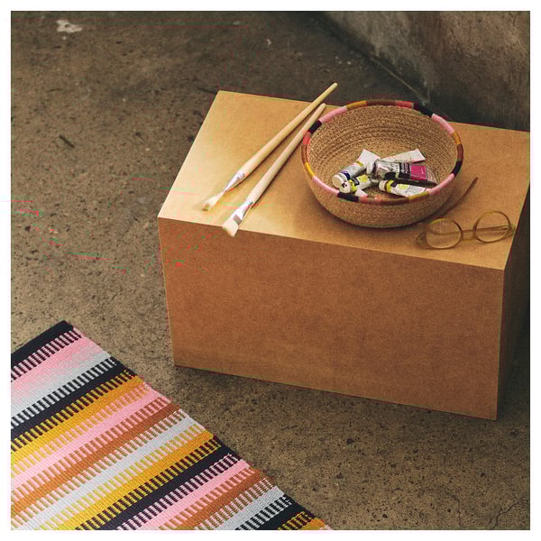 Jute MÄVINN basket with art supplies on cork cube, colorful rug, round glasses.