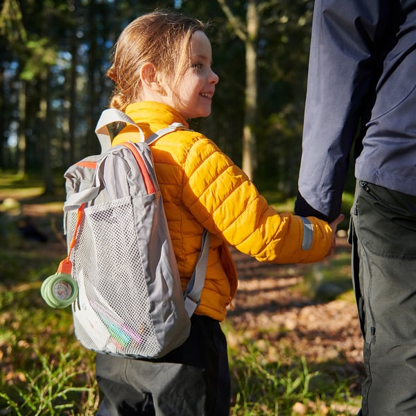 A child wearing a yellow jacket carries a light grey MAJBAGGE backpack with mesh pockets and a compass.