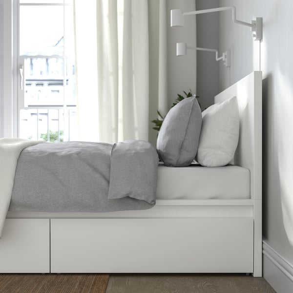 Modern white MALM bed with storage, grey bedding, plant, and wall lamp near window.