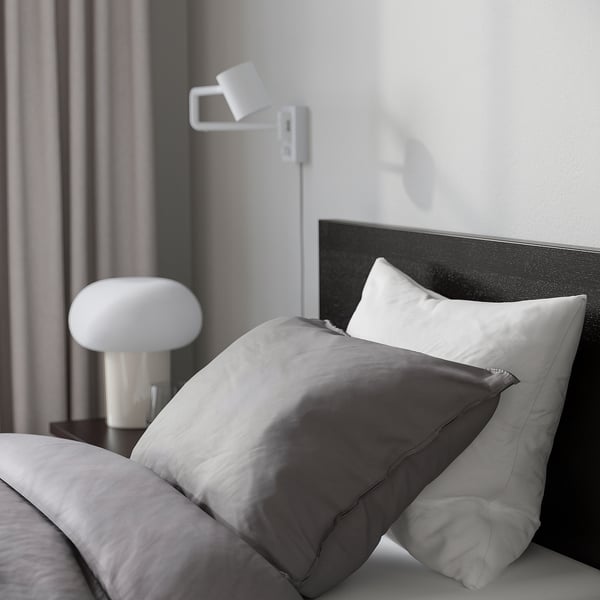 Bedroom scene with modern minimalist design. Grey bed with high headboard, grey pillows, side lamp, and wall-mounted light.