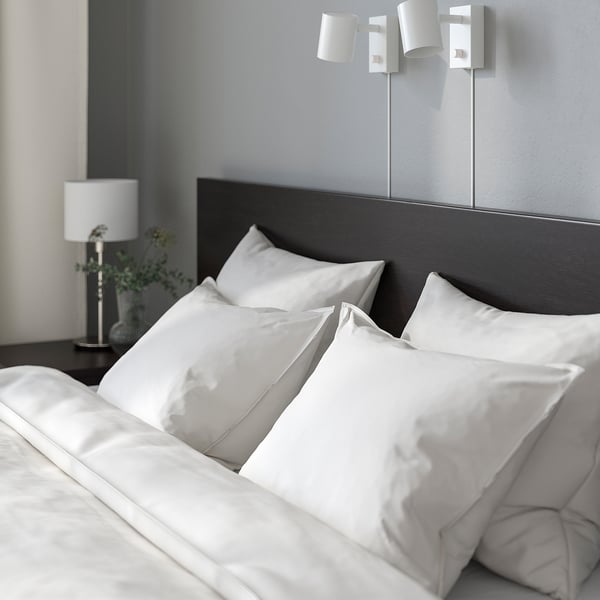 Bedroom scene with white bed, dark headboard, wall lamps, and bedside table with lamp and plant.