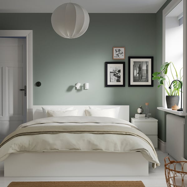 Bedroom with white MALM bed, light green walls, hanging light, black framed photos, plant, and wicker chair.