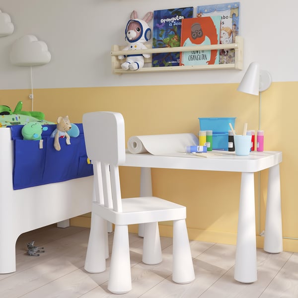 Small childs white table & chair set with storage pockets, placed under a shelf of colourful books. Ideal for play & crafts.