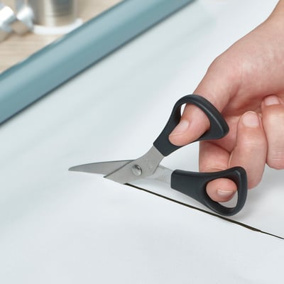 Hand holding gray-handled scissors cutting white paper on a light surface.