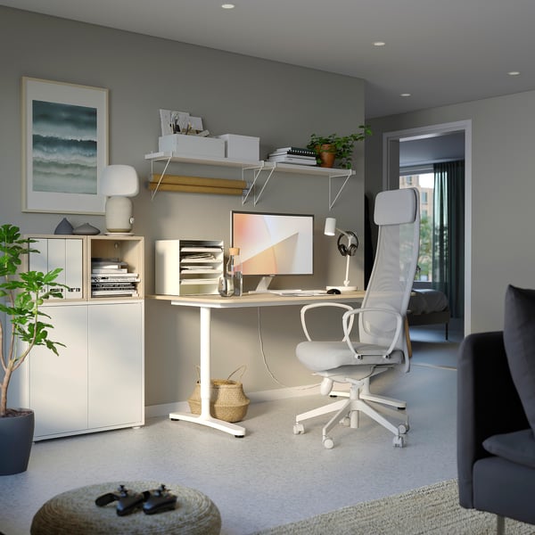 Modern home office with white ergonomic chair, wooden desk, shelves, computer, lamp, and plants.
