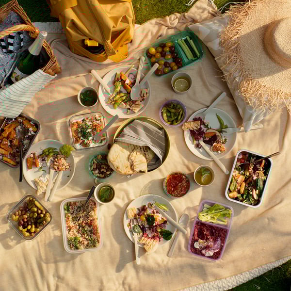 Picnic spread with MARULK beige set on blanket. Includes plates, cutlery, trays, and containers with food. Yellow bag and a wine bottle nearby.