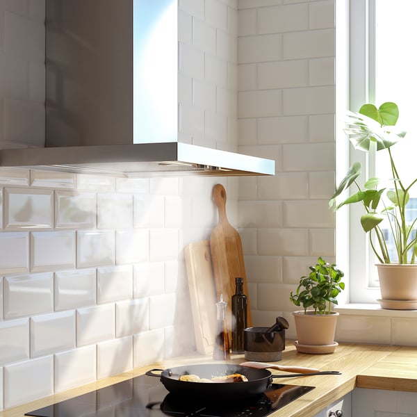 Modern kitchen with white brick backsplash and stainless steel hood. Wooden utensils, plants, and cooking pan on the countertop.