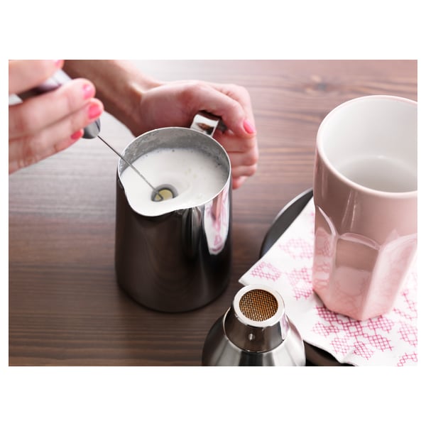 Person frothing milk in a metal jug on a wooden table, next to a pink mug and a metal strainer.