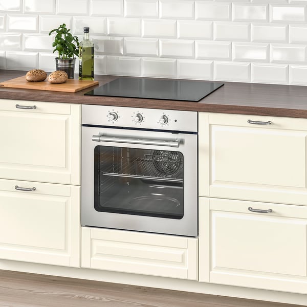 Modern kitchen with silver oven, white cabinets, black cooktop. Counter has bread, plant, olive oil. Brick-style backsplash.