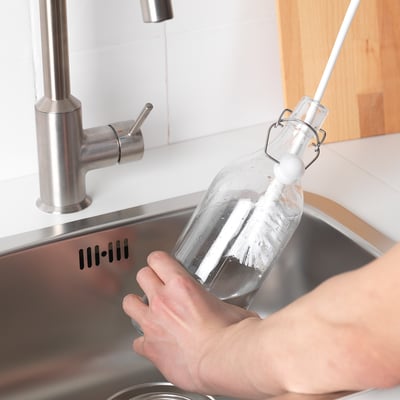 Person holding a clear bottle with a long-handled brush under a tap.