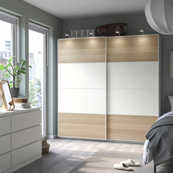Modern bedroom with light-brown & white MEHAMN sliding closet, wooden floor, grey walls, large window, cosy rug, and bed.
