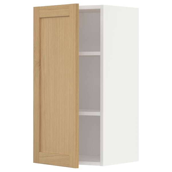 Displays a white METOD kitchen wall cabinet with a single open wooden door, featuring oak veneer and beveled edges.