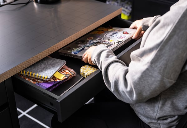 Person in grey hoodie reaching into black MICKE desk drawer with colorful notebooks, a yellow object, and cables.