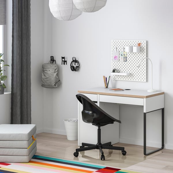 Modern home office with a white MICKE desk, black chair, and colorful rug. Pegboard with accessories and decorative lanterns.