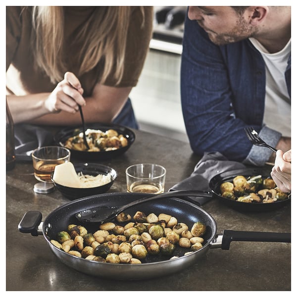 Two people eat roasted brussels sprouts from black bowls and pans on a dark table, with glasses of beer.