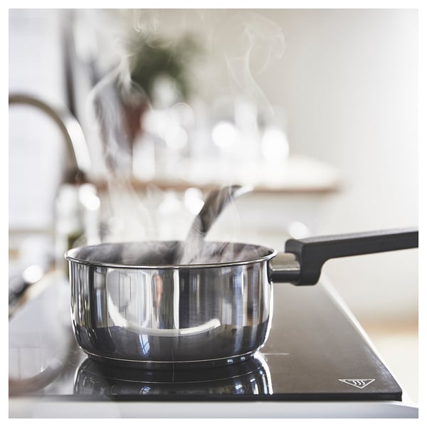 Displays a MIDDAGSMAT saucepan on a black induction hob, emitting steam. The pan is silver with a black handle, featuring a sleek, modern design.