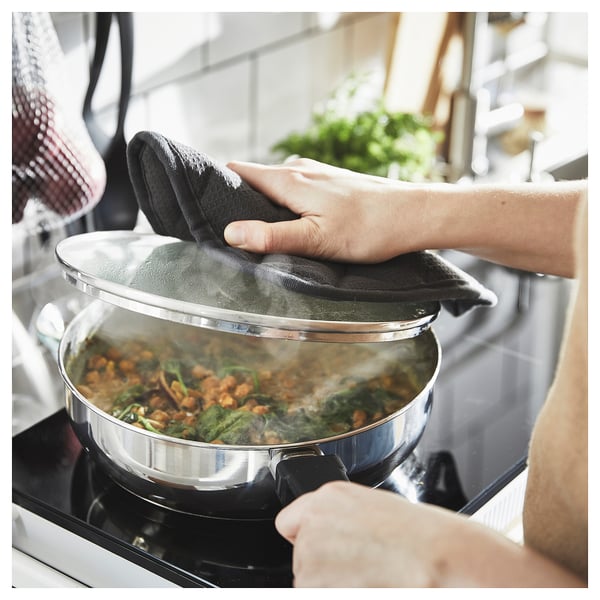 A person lifts a steaming sauté pan on a stove, wearing black oven mitts.