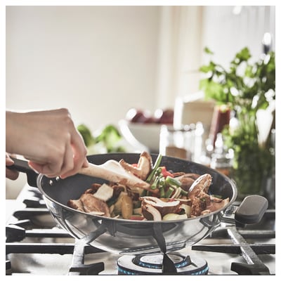 Person cooking in a clear glass wok on a stove. Ingredients visible inside. Kitchen setting with green plants.