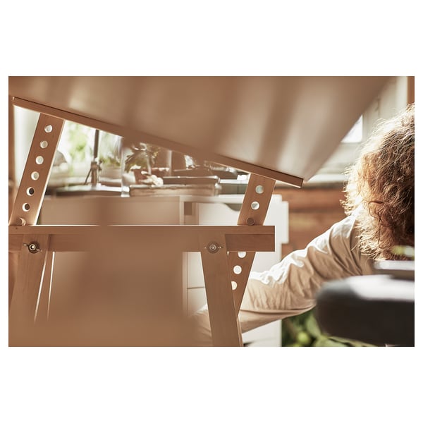A person adjusts a wooden MITTBACK trestle table, featuring solid birch legs with tilt function, ideal for various tasks.