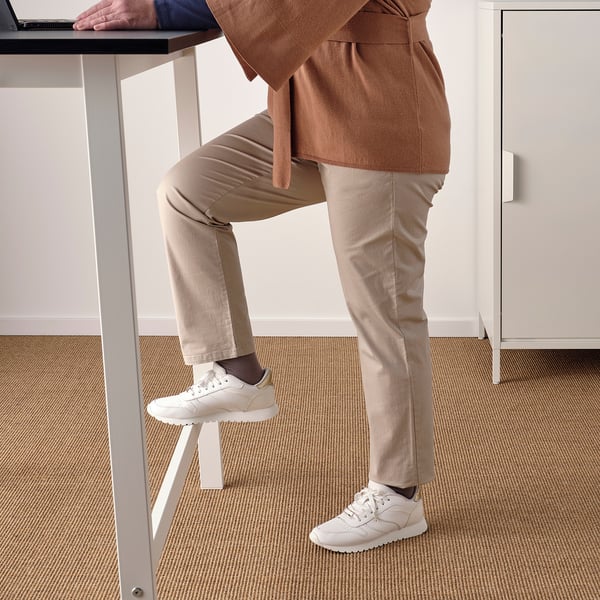 Person leaning on standing desk with feet on stool, wearing white trainers and beige pants.