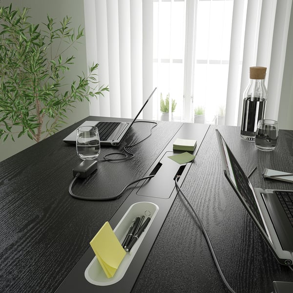 Modern conference table with built-in slots for devices, pens, and notepads, equipped with power outlets, near a window with plants.