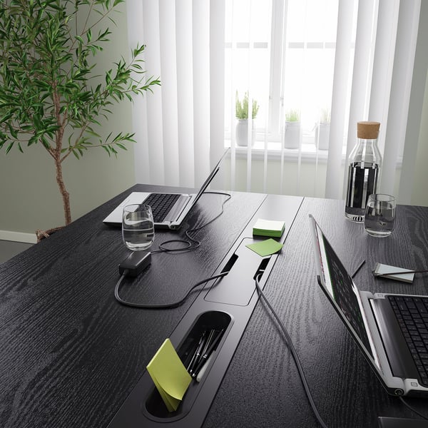 Modern black conference table with integrated power outlets and cable management, holding laptops, cups, and a carafe.