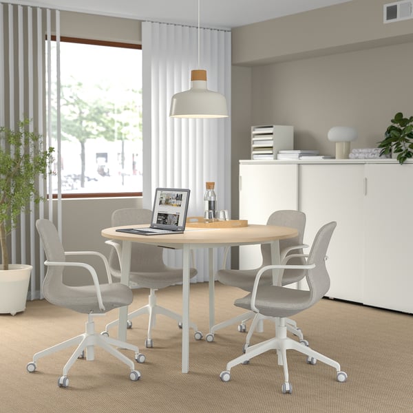 Modern meeting space with round table, chairs, laptop, lamp. Bright room with large window, beige decor.