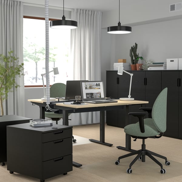 Modern office with adjustable desk, green chairs, computer, lights, printer, plants, large window.