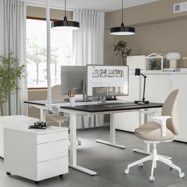 Modern office setup featuring MITTZON white standing desk with black top, ergonomic chair, dual pendant lights, and a monitor. Tall, sleek design.
